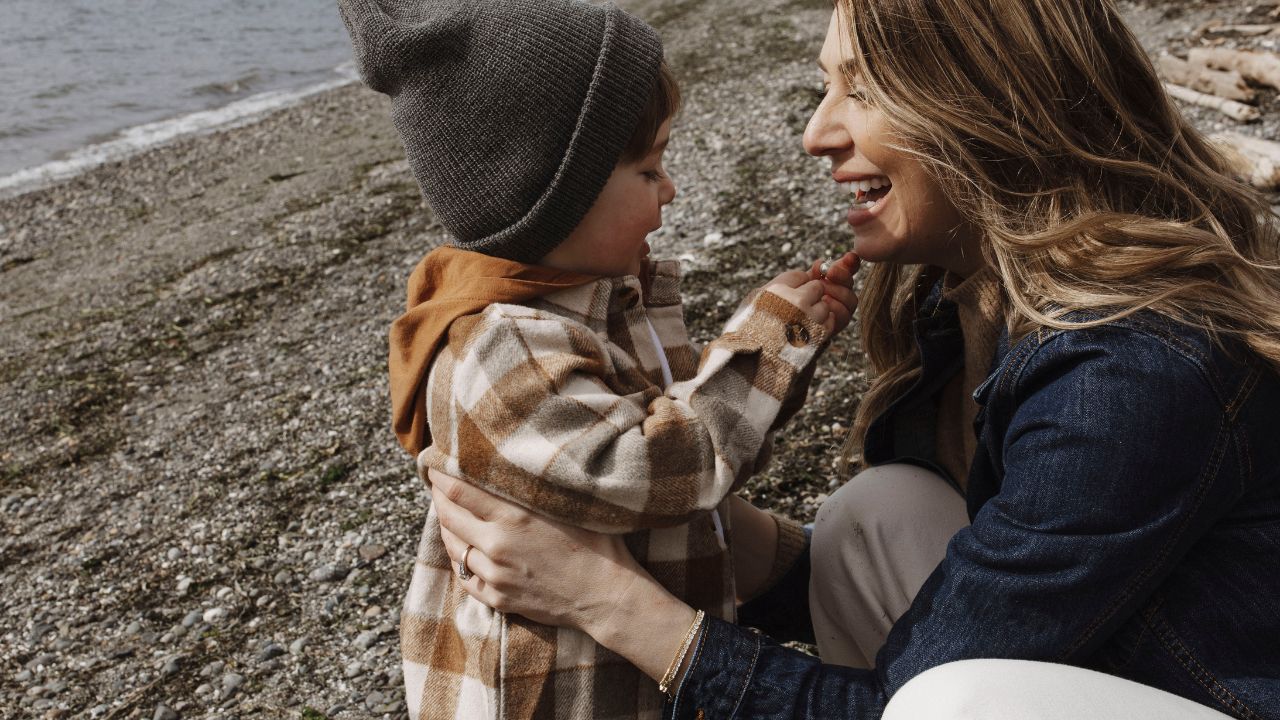 Mother smiling and kneeling beside her young child on a rocky beach, both dressed in cozy fall clothing; the child wears a gray beanie and plaid jacket while touching the mother’s face, capturing a warm, candid moment of connection.